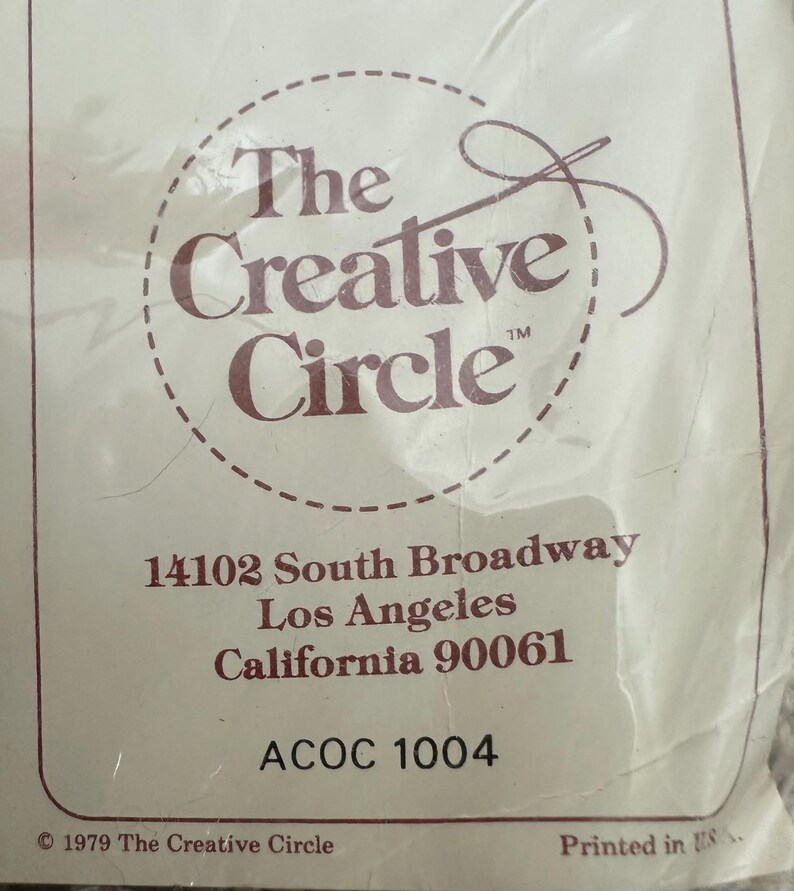 May include: A white label with brown text that reads "The Creative Circle" with a stylized needle and thread logo. The label also includes the address "14102 South Broadway, Los Angeles, California 90061" and the code "ACOC 1004". The bottom of the label reads "&copy; 1979 The Creative Circle" and "Printed in USA."