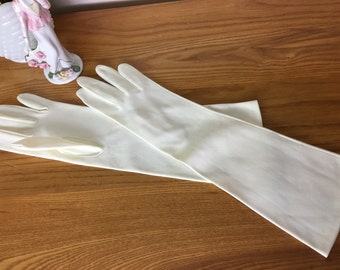 May include: A pair of long, white gloves made of a smooth fabric. The gloves are laid out flat on a wooden surface.