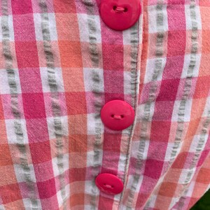 K&ouml;nnte beinhalten: Ein rosa-wei&szlig; kariertes Kleid mit vier gro&szlig;en rosa Kn&ouml;pfen auf dem R&uuml;cken.