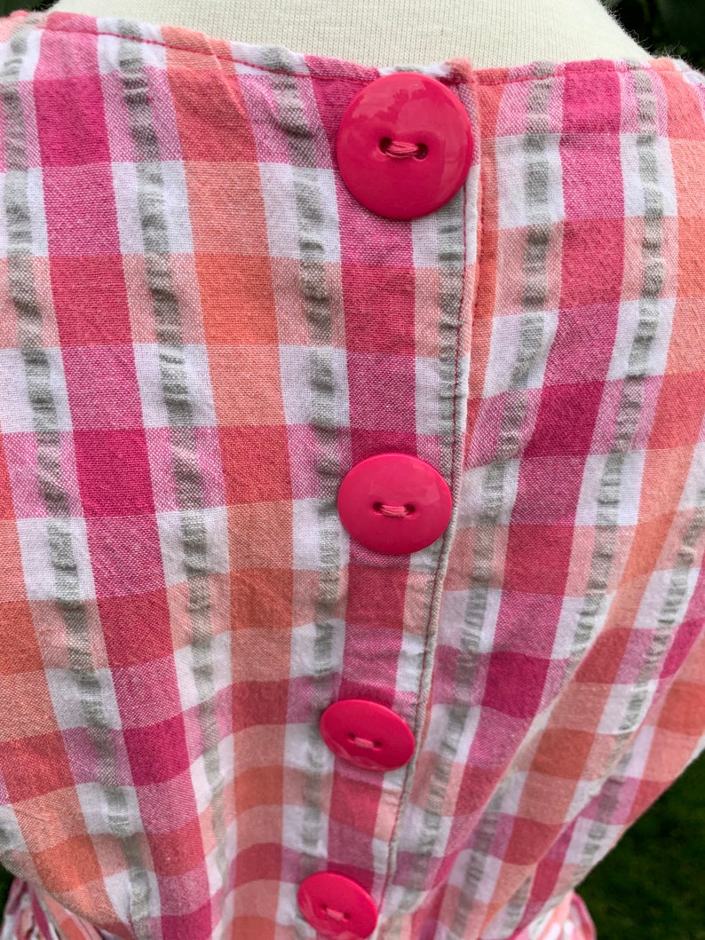 K&ouml;nnte beinhalten: Ein rosa-wei&szlig; kariertes Kleid mit vier gro&szlig;en rosa Kn&ouml;pfen auf dem R&uuml;cken.