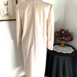 K&ouml;nnte beinhalten: Ein langer, cremefarbener Mantel mit einem schwarz-wei&szlig;en Kragen. Der Mantel befindet sich auf einem Mannequin und wird von hinten fotografiert.
