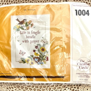 May include: A yellow and white embroidery kit with a bird, flowers, and the text "Life is fragile handle with prayer". The kit is from The Creative Circle and has the product number 1004.
