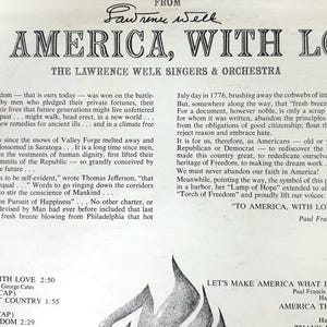 Op de afbeelding: Een achterkant van een vintage vinylplaat met de titel TO AMERICA, WITH LOVE door The Lawrence Welk Singers & Orchestra. De witte papieren hoes bevat zwarte gedrukte tekst, waaronder een lang essay van Paul Francis Webster en een tracklist voor zijde 1 en 2.