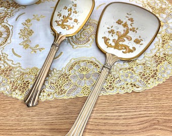 May include: Two hand mirrors with long, fluted gold-tone metal handles and rectangular heads. The back of each mirror features a cream-coloured surface decorated with an ornate gold and pink floral pattern, showcasing intricate scrollwork and small blossoms.