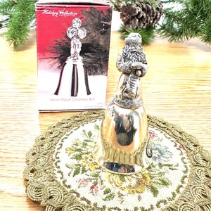 Pode incluir: Um sino de Natal prateado com uma figura do Papai Noel. O sino est&aacute; sobre uma toalha de mesa floral com uma borda dourada. Uma caixa com o texto "Holiday Collection" e uma imagem de sino est&aacute; no fundo.