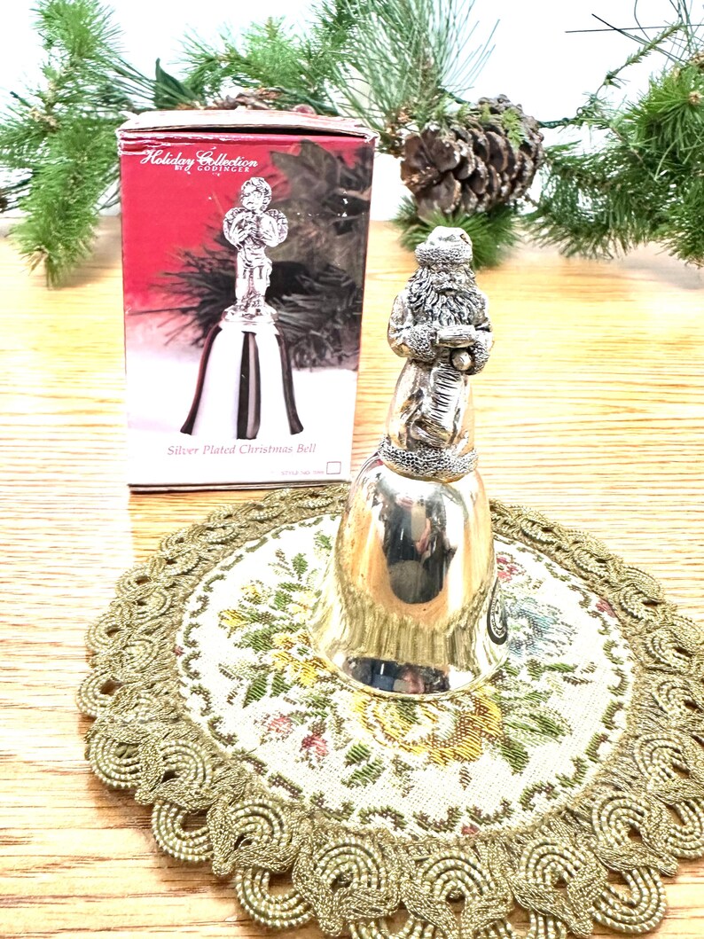 Pode incluir: Um sino de Natal prateado com uma figura do Papai Noel. O sino est&aacute; sobre uma toalha de mesa floral com uma borda dourada. Uma caixa com o texto "Holiday Collection" e uma imagem de sino est&aacute; no fundo.
