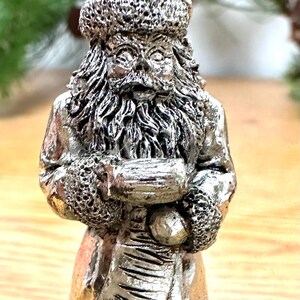 Pode incluir: Estatueta prateada do Pai Natal. A figura tem um chap&eacute;u, barba e casaco texturizados, segurando um saco. &Eacute; uma decora&ccedil;&atilde;o festiva, perfeita para a &eacute;poca natal&iacute;cia.