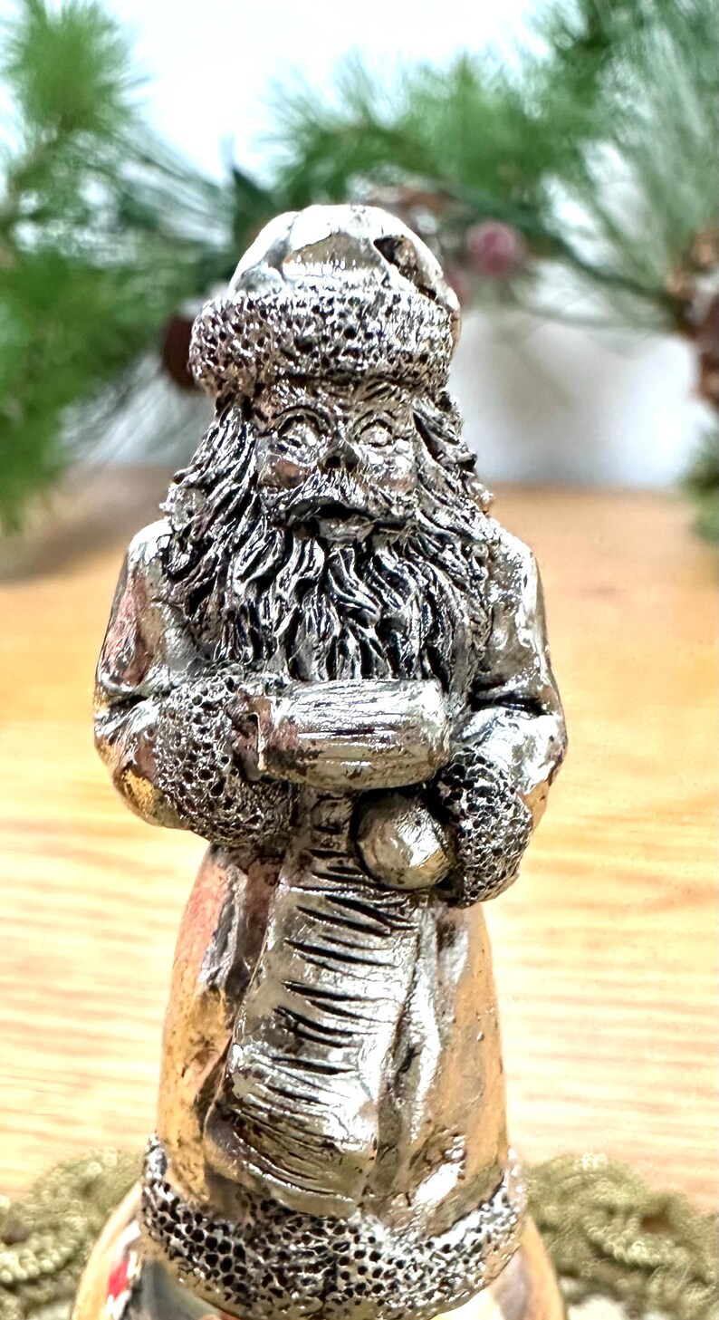 Pode incluir: Estatueta prateada do Pai Natal. A figura tem um chap&eacute;u, barba e casaco texturizados, segurando um saco. &Eacute; uma decora&ccedil;&atilde;o festiva, perfeita para a &eacute;poca natal&iacute;cia.