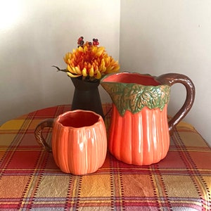 May include: A set of orange pumpkin-shaped ceramic kitchenware, including a pitcher and a mug. The pitcher has a brown handle and green leaves around the top. The mug has a brown handle. The items are on a red and yellow plaid tablecloth.