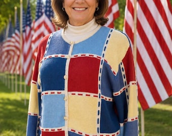 May include: A colorful patchwork cardigan in red, white, blue, and yellow. The sweater has a button-up front and is worn over a white turtleneck. American flags are in the background.