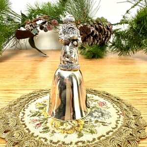 Pode incluir: Um sino prateado com uma figura do Pai Natal no topo, colocado sobre uma toalha de mesa floral com acabamento dourado. O sino &eacute; rodeado por ramos de pinheiro e uma pinha, sugerindo um tema de f&eacute;rias.