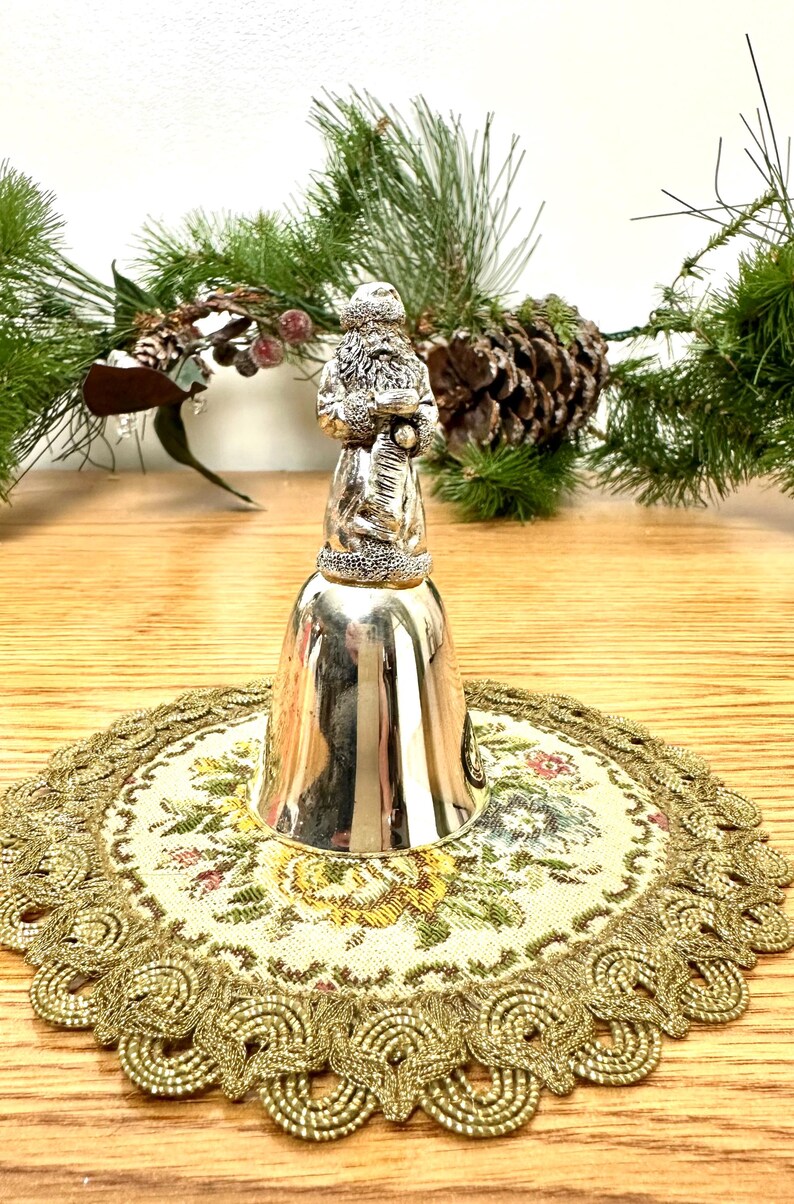Pode incluir: Um sino prateado com uma figura do Pai Natal no topo, colocado sobre uma toalha de mesa floral com acabamento dourado. O sino &eacute; rodeado por ramos de pinheiro e uma pinha, sugerindo um tema de f&eacute;rias.