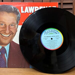 Op de afbeelding: Een zwarte vinylplaat met een Ranwood-label getiteld To America With Love, voorzien van een tracklist en een middengat. Daarnaast ligt een platenhoes met een portret van Lawrence Welk in een bruin pak, lichtblauw overhemd en een das met patroon.