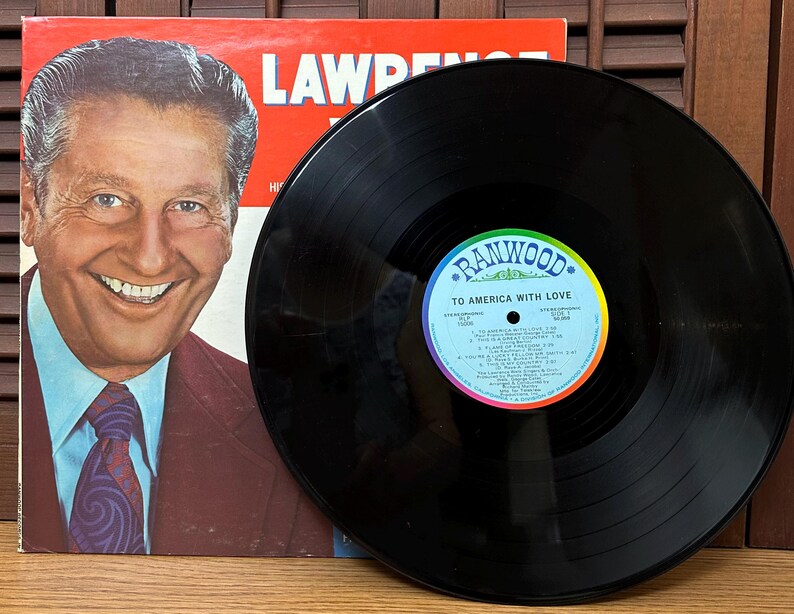 Op de afbeelding: Een zwarte vinylplaat met een Ranwood-label getiteld To America With Love, voorzien van een tracklist en een middengat. Daarnaast ligt een platenhoes met een portret van Lawrence Welk in een bruin pak, lichtblauw overhemd en een das met patroon.