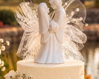 May include: A white ceramic wedding cake topper featuring a couple standing face to face, adorned with a white tulle fan backdrop accented by small silver-tipped pearl beads, positioned atop a frosted white cake decorated with white pearl dots.
