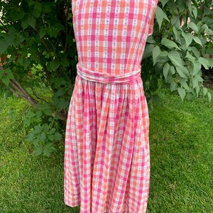 K&ouml;nnte beinhalten: Ein rosa-oranges Gingham-Kleid ohne &Auml;rmel mit einer wei&szlig;en Sch&auml;rpe. Das Kleid hat einen vollen Rock und ist aus einem leichten Stoff gefertigt.