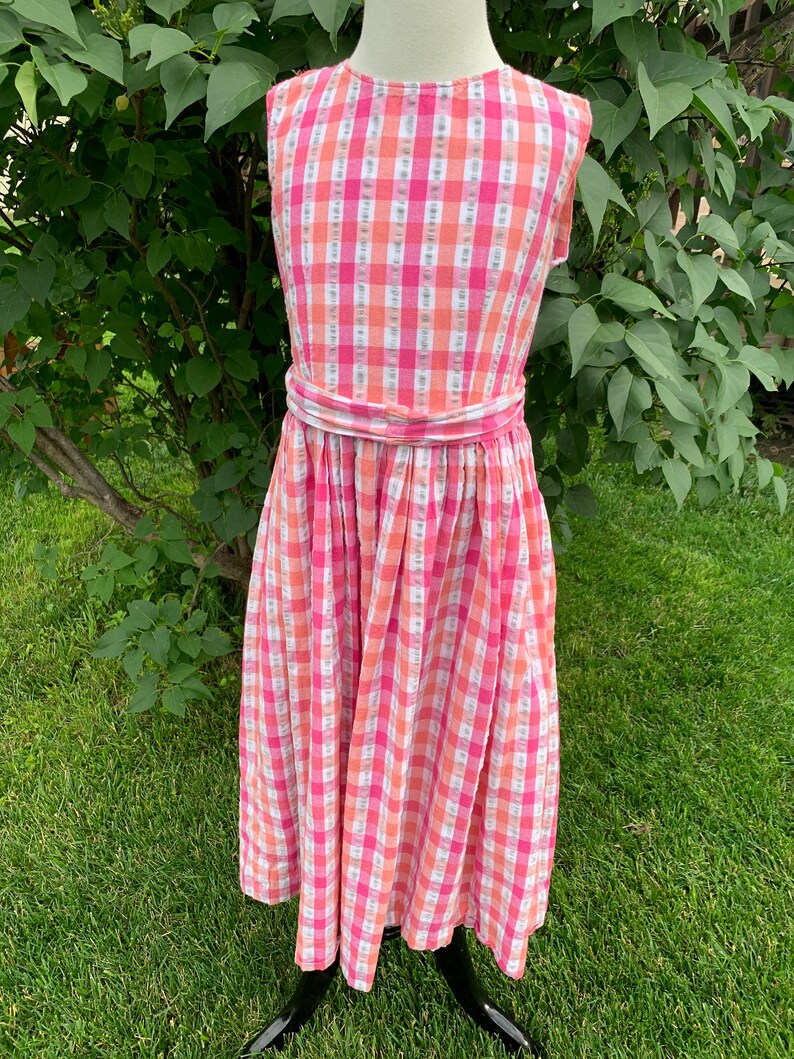 K&ouml;nnte beinhalten: Ein rosa-oranges Gingham-Kleid ohne &Auml;rmel mit einer wei&szlig;en Sch&auml;rpe. Das Kleid hat einen vollen Rock und ist aus einem leichten Stoff gefertigt.