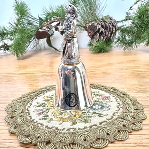Pode incluir: Um sino prateado com uma figura do Pai Natal no topo. O sino est&aacute; sobre uma toalha de mesa redonda com um padr&atilde;o floral e uma borda dourada e ondulada. O fundo inclui ramos de pinheiro e uma pinha.