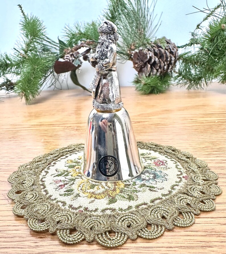 Pode incluir: Um sino prateado com uma figura do Pai Natal no topo. O sino est&aacute; sobre uma toalha de mesa redonda com um padr&atilde;o floral e uma borda dourada e ondulada. O fundo inclui ramos de pinheiro e uma pinha.