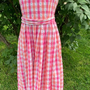 K&ouml;nnte beinhalten: Ein rosa-oranges Gingham-Kleid ohne &Auml;rmel mit einem passenden G&uuml;rtel. Das Kleid befindet sich auf einer Puppe und wird vor einem gr&uuml;nen Gras Hintergrund pr&auml;sentiert.