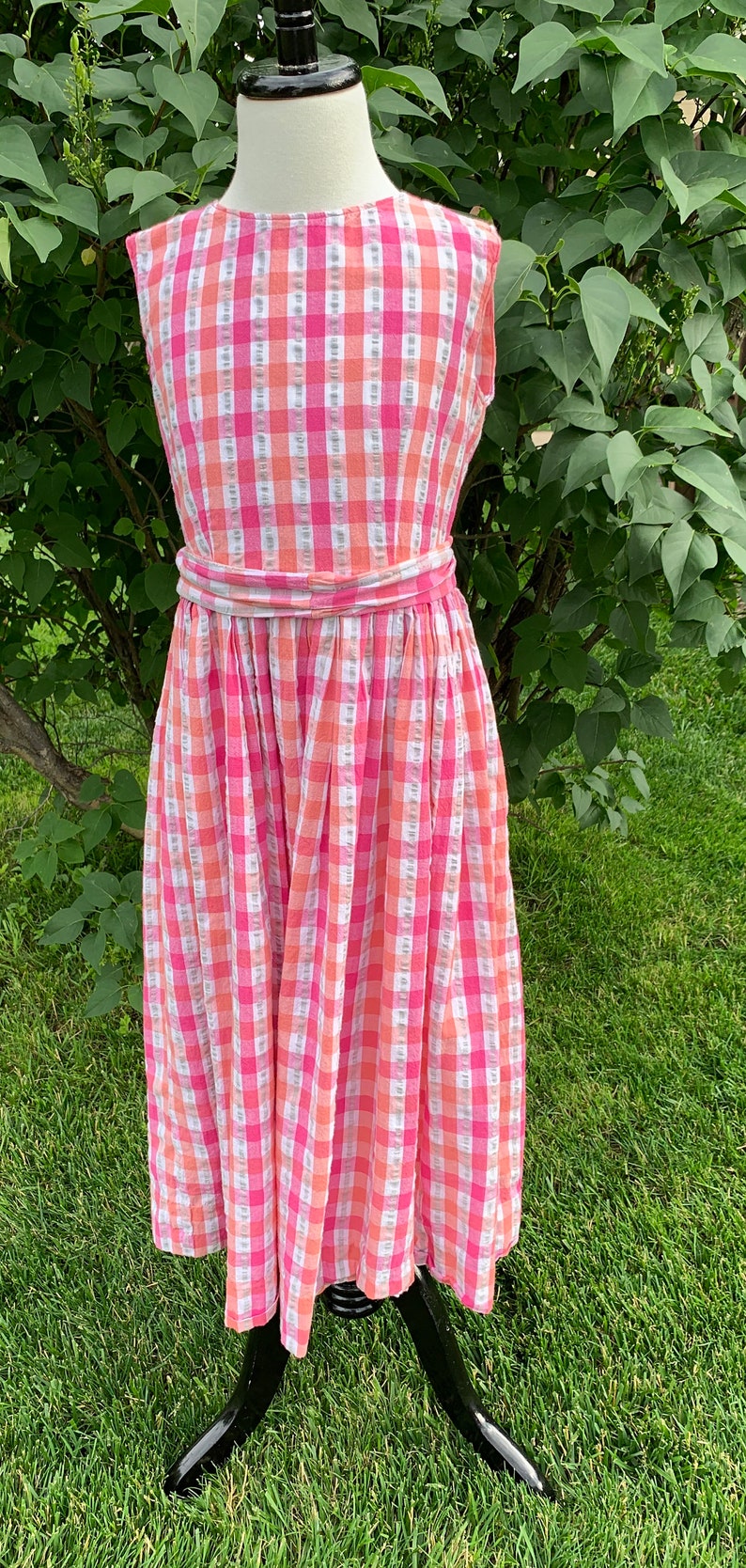 K&ouml;nnte beinhalten: Ein rosa-oranges Gingham-Kleid ohne &Auml;rmel mit einem passenden G&uuml;rtel. Das Kleid befindet sich auf einer Puppe und wird vor einem gr&uuml;nen Gras Hintergrund pr&auml;sentiert.