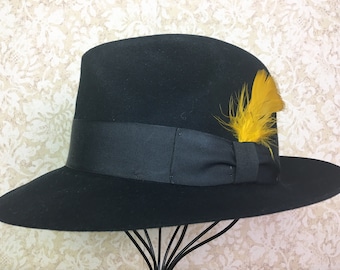 May include: A black fedora hat with a black ribbon and a yellow feather accent.