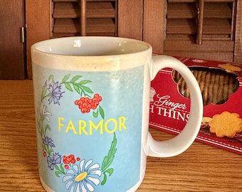 May include: A white ceramic mug featuring a light blue rectangular panel with a floral wreath design composed of blue cornflowers, white daisies, and red berries. The word FARMOR is printed in yellow capital letters in the center of the wreath.