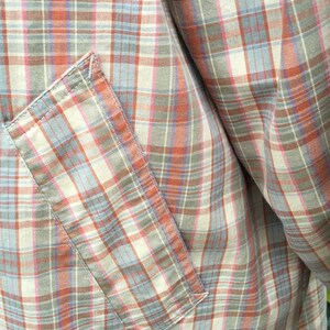 May include: A close-up of a plaid shirt with a patch pocket. The shirt is made of a soft, lightweight fabric and features a multi-colored plaid pattern in shades of blue, pink, and brown.