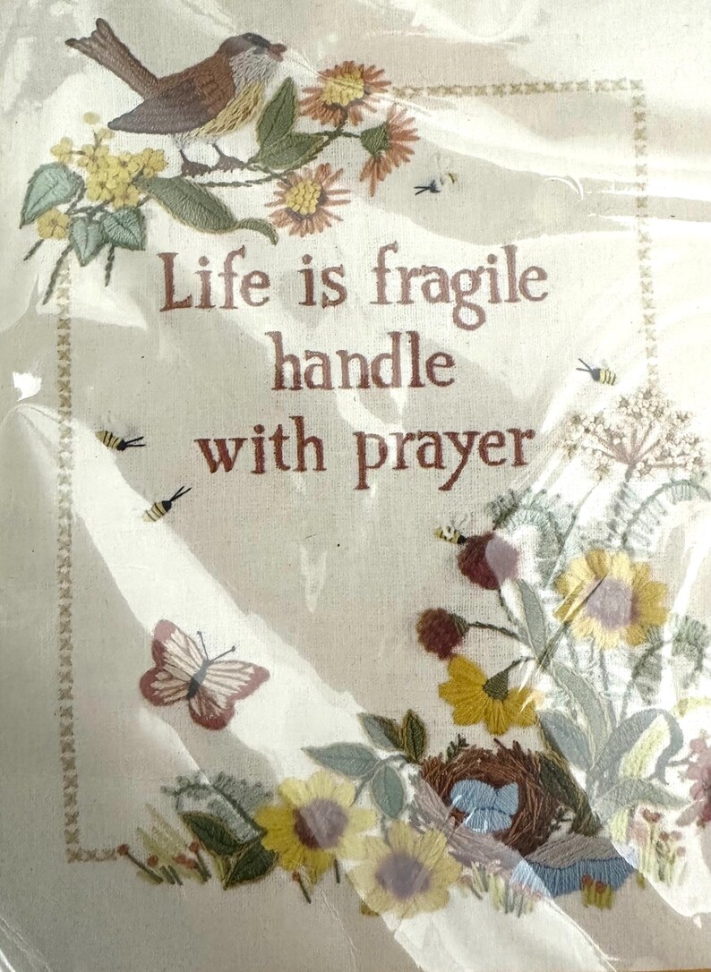 May include: Embroidered wall art with a bird, flowers, and butterflies. The text reads "Life is fragile handle with prayer".