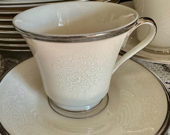 May include: A cream-coloured porcelain teacup and saucer set featuring a delicate white embossed floral pattern and a polished silver rim. The teacup has a curved handle and sits centred on the matching saucer, which displays a matching floral border.