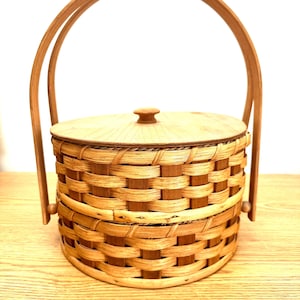 May include: A round, woven wicker basket with a lid and two wooden handles. The basket is brown and has a natural finish.