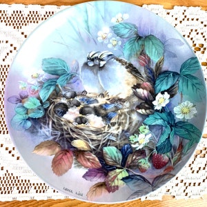 May include: A blue and white porcelain plate featuring a painted image of a bird family in a nest. The birds are surrounded by green leaves and white flowers. The plate is signed "Lena Lilj" in the bottom right corner.
