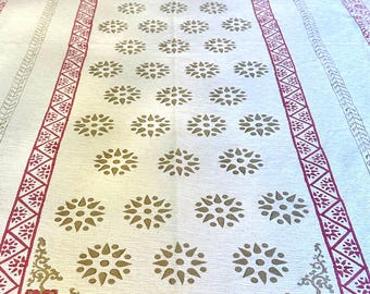 May include: Cream-colored tablecloth with a repeating floral pattern in gold and a red border. The border features geometric and floral designs. The tablecloth is rectangular and covers a wooden table.