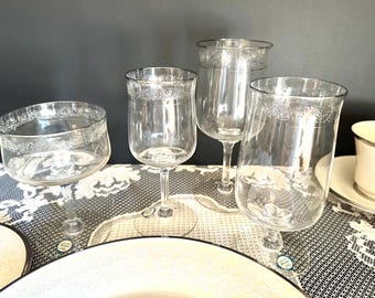 May include: A collection of four clear glass stemware pieces featuring a delicate etched floral band near the rim. The set includes a wide-mouthed coupe glass, two varying sizes of stemmed wine glasses, and a tall, slender glass, all with clear stems.