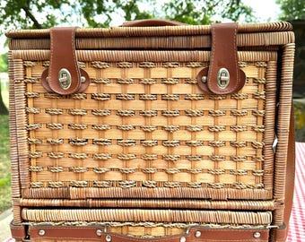 May include: A woven wicker picnic basket with a brown leather handle and straps. The basket has a rectangular shape with a hinged lid and a lower compartment. The basket is sitting on a red and white checkered tablecloth.