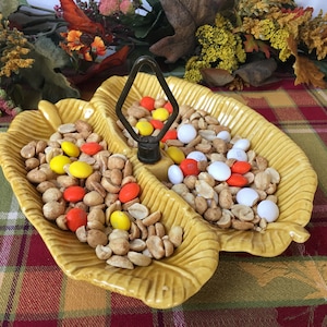 May include: A yellow ceramic serving dish shaped like two leaves with a metal divider in the center. The dish is filled with peanuts and candy.