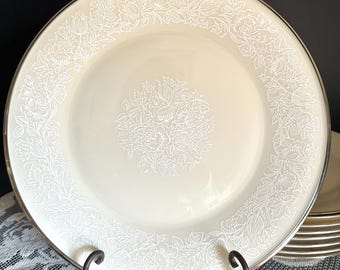 May include: A round cream-coloured dinner plate featuring a silver rim and a white embossed floral pattern. The design includes a central circular cluster of flowers and leaves, surrounded by a continuous border of matching floral motifs.