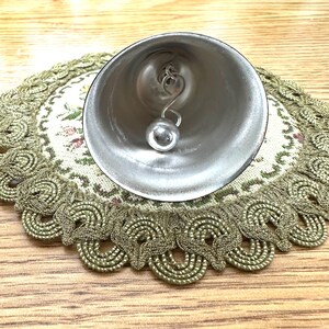 Pode incluir: Um sino prateado com um badalo esf&eacute;rico pendurado no interior. O sino est&aacute; sobre uma toalha de mesa decorativa com um padr&atilde;o floral e uma borda ondulada dourada. O fundo &eacute; uma superf&iacute;cie de madeira clara.