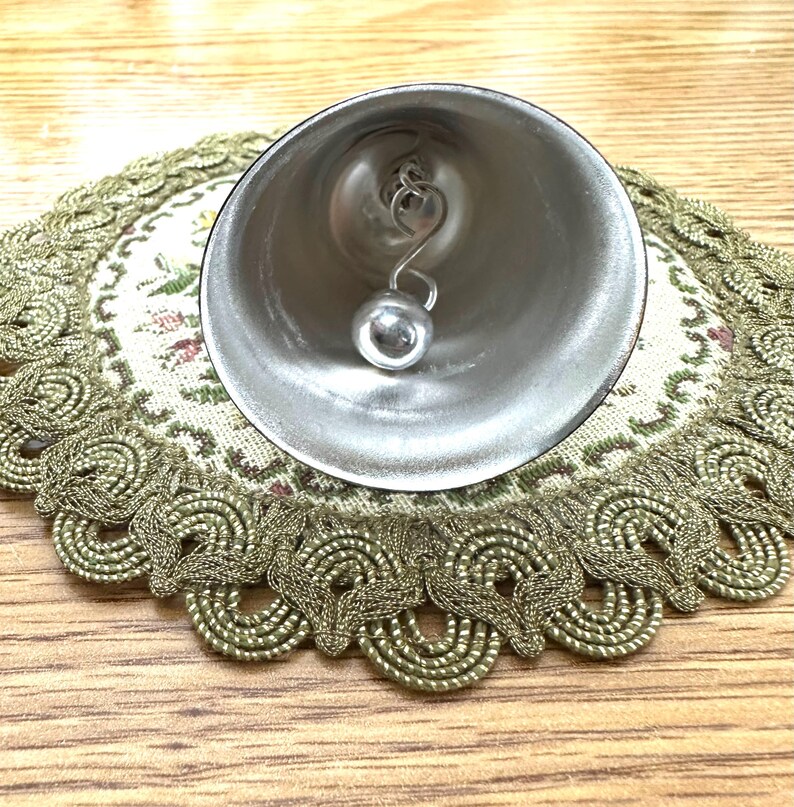 Pode incluir: Um sino prateado com um badalo esf&eacute;rico pendurado no interior. O sino est&aacute; sobre uma toalha de mesa decorativa com um padr&atilde;o floral e uma borda ondulada dourada. O fundo &eacute; uma superf&iacute;cie de madeira clara.
