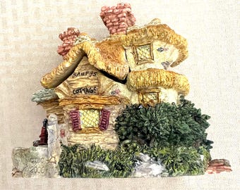 May include: A small, detailed, ceramic cottage figurine with a thatched roof, a sign that reads "Bailey's Cottage", and a green bush. The base of the figurine is decorated with a stone-like texture and the year 2000.