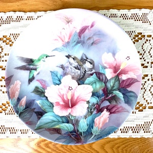 May include: A decorative plate with a hummingbird and flowers. The plate features a pink hibiscus flower, green leaves, and a hummingbird in flight. The artist's signature, "Lena Liu", is visible in the bottom right corner.