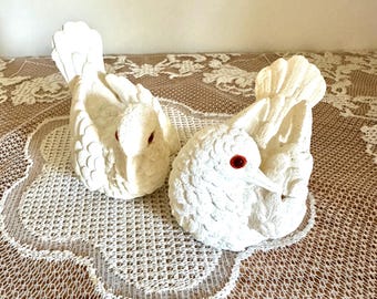 May include: Two white bird figurines with textured feather details and small, round, bright red eyes. Each bird is depicted in a resting position with wings folded against its body and a fanned tail, crafted from a matte, porous material.