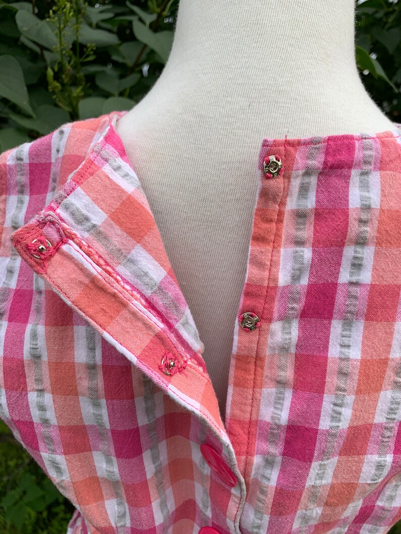 K&ouml;nnte beinhalten: Ein rosa- und orangefarbenes kariertes Hemd mit Kn&ouml;pfen auf wei&szlig;em Grund. Das Hemd hat einen Knopfverschluss und einen Kragen.
