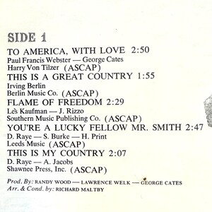Op de afbeelding: Een tracklist op wit papier met de titel SIDE 1 en zwarte tekst. De nummers zijn onder andere TO AMERICA, WITH LOVE, THIS IS A GREAT COUNTRY, FLAME OF FREEDOM, YOU'RE A LUCKY FELLOW MR. SMITH en THIS IS MY COUNTRY met bijbehorende credits.