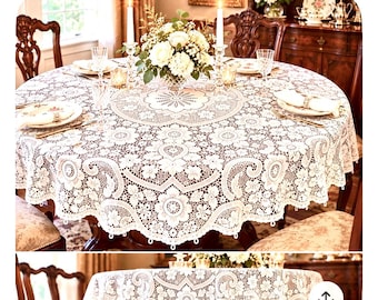 May include: A round white lace tablecloth featuring an intricate floral pattern with scalloped edges. The fabric displays a detailed openwork design with circular motifs and botanical elements, draping over a circular dining table surface.