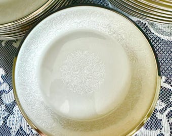 May include: A stack of off-white ceramic plates featuring a central embossed floral motif and a matching floral pattern along the rim. Each plate is finished with a metallic silver band around the edge, creating a reflective border on the dinnerware.