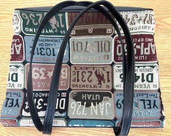 May include: A rectangular tote bag featuring a woven tapestry pattern of various vintage-style number plates in shades of blue, green, red, and brown. The bag includes two black shoulder straps and a solid black top trim border.