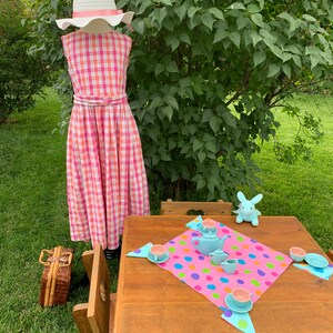 K&ouml;nnte beinhalten: Ein rosa-wei&szlig; kariertes Kleid mit einem wei&szlig;en Hut, ein Holztisch mit einer rosa Tischdecke mit bunten Punkten, eine blaue Teekanne und Tassen sowie ein blaues Hasenspielzeug.