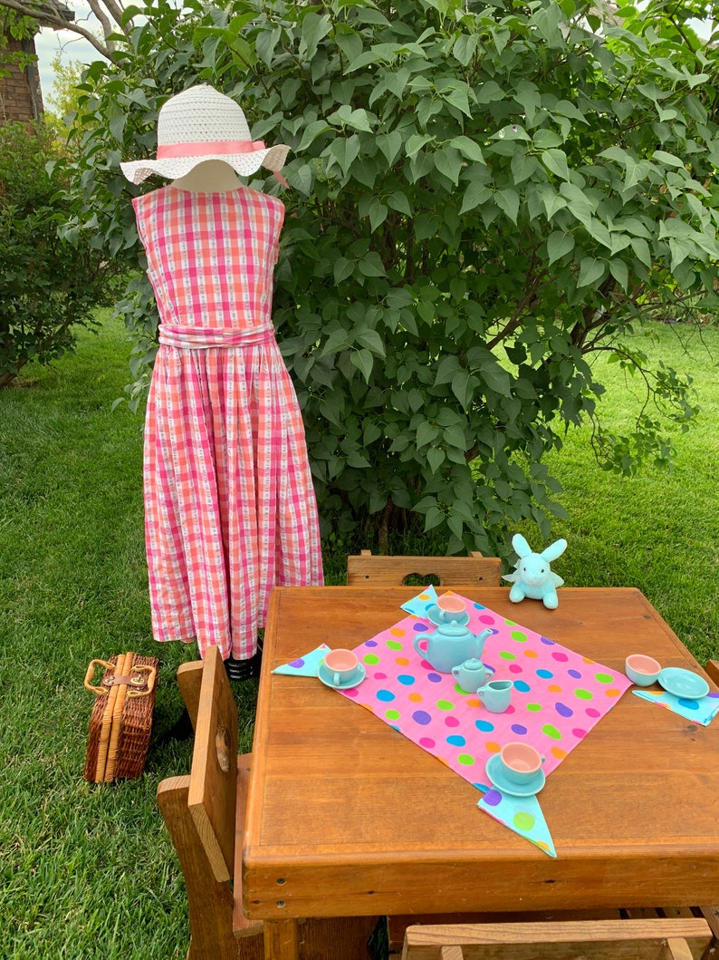 K&ouml;nnte beinhalten: Ein rosa-wei&szlig; kariertes Kleid mit einem wei&szlig;en Hut, ein Holztisch mit einer rosa Tischdecke mit bunten Punkten, eine blaue Teekanne und Tassen sowie ein blaues Hasenspielzeug.