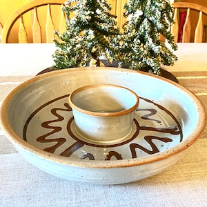May include: A speckled, off-white ceramic chip and dip serving dish with a central dip bowl. The dish features brown abstract designs and a brown rim. Two small, snow-covered Christmas trees are in the background.
